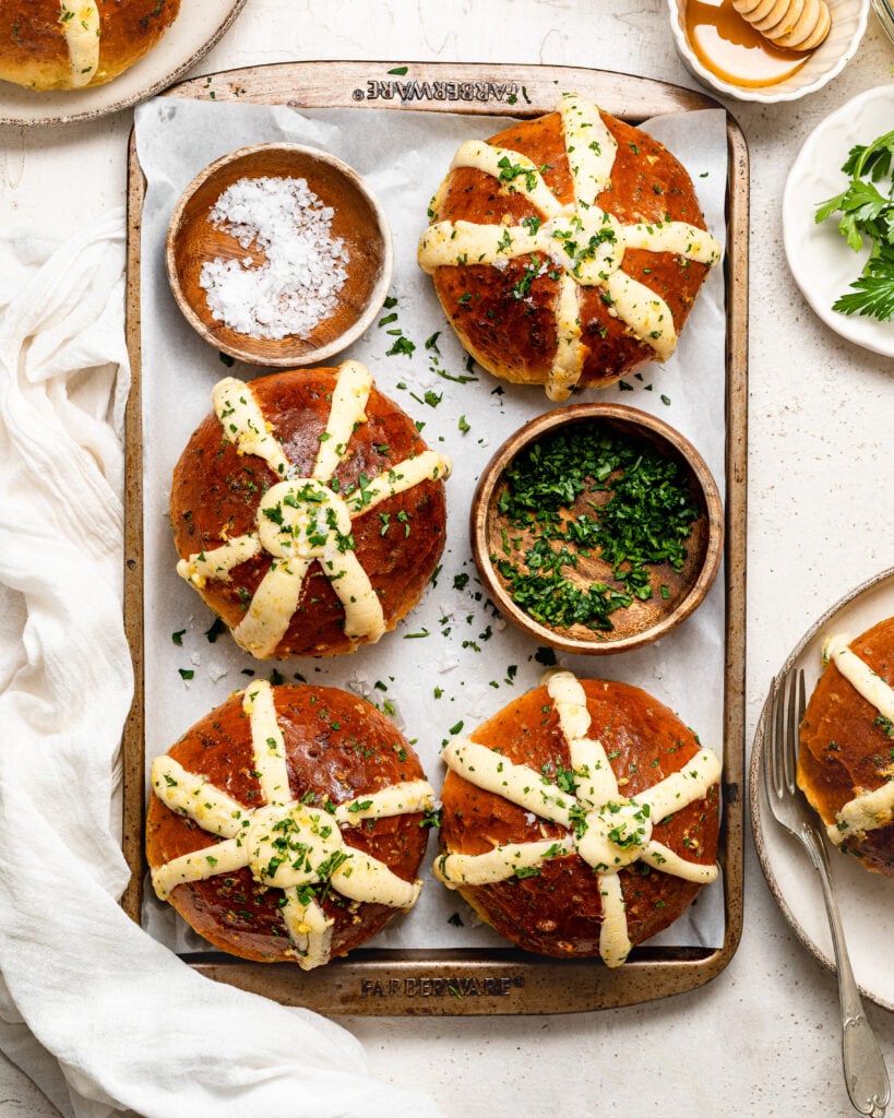Golden Korean cream cheese garlic bread buns with pale filling and chopped herbs on a tray - Catherine Zhang Three pretzel buns on a parchment-lined tray, brushed with herb butter crosses and chopped parsley.