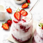Glass of strawberry milkshake topped with sliced strawberries and ice on a marble surface
