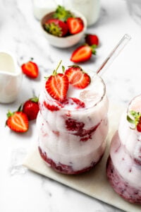 Glass of strawberry milkshake topped with sliced strawberries and ice on a marble surface
