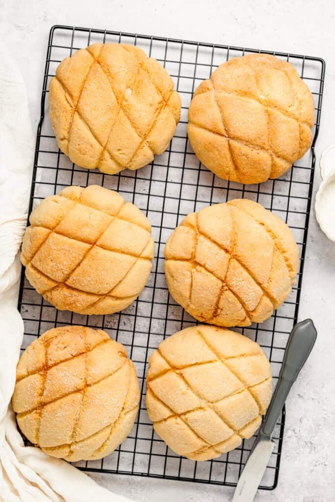 Round melon-pan buns with a grid pattern cooling on a wire rack over a light countertop, with a knife nearby