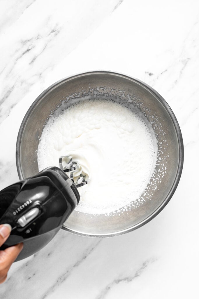 Hand mixer whipping a foamy white cream mixture in a metal bowl for croffles - Catherine Zhang Hand mixer whipping a foamy white cream mixture in a metal bowl for croffles
