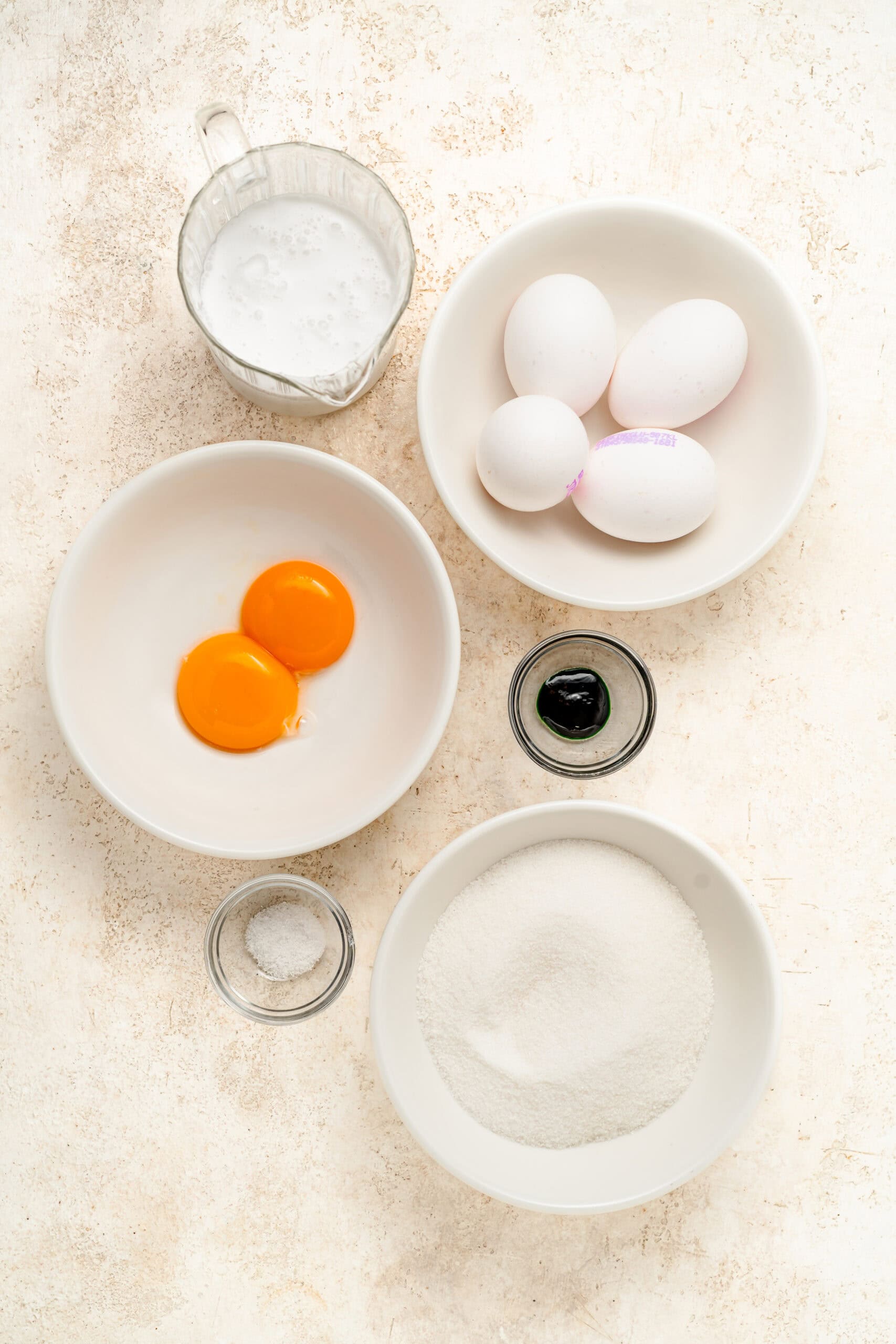 Ingredients - Pandan Kaya Jam - Catherine Zhang Top-down view of baking ingredients: eggs in bowls, milk in a pitcher, vanilla extract, and a bowl of sugar on a light countertop.