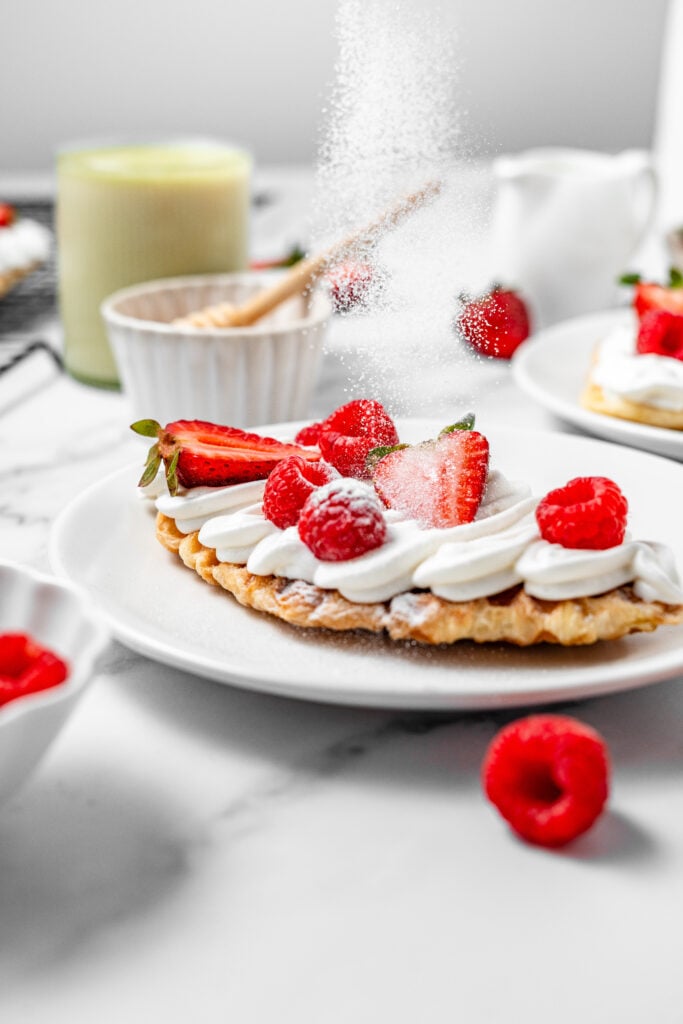 Powdered sugar falling over a plated croffle topped with whipped cream and berries - Catherine Zhang Powdered sugar falling over a plated croffle topped with whipped cream and berries