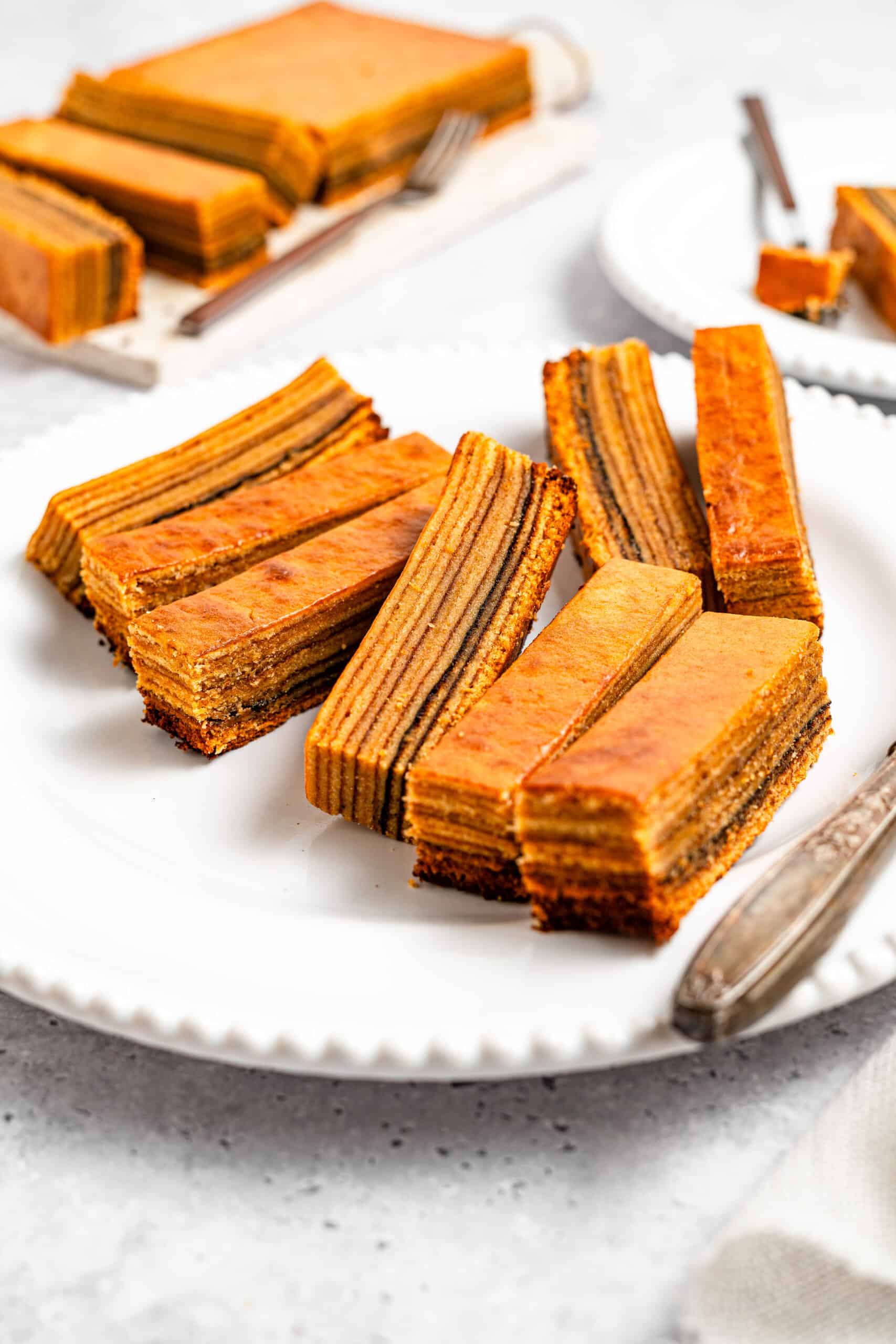 Stacked slices of lapis legit showing even caramelised layers - Catherine Zhang Slices of layered honey cake with orange glaze arranged on a white plate on a rustic marble surface