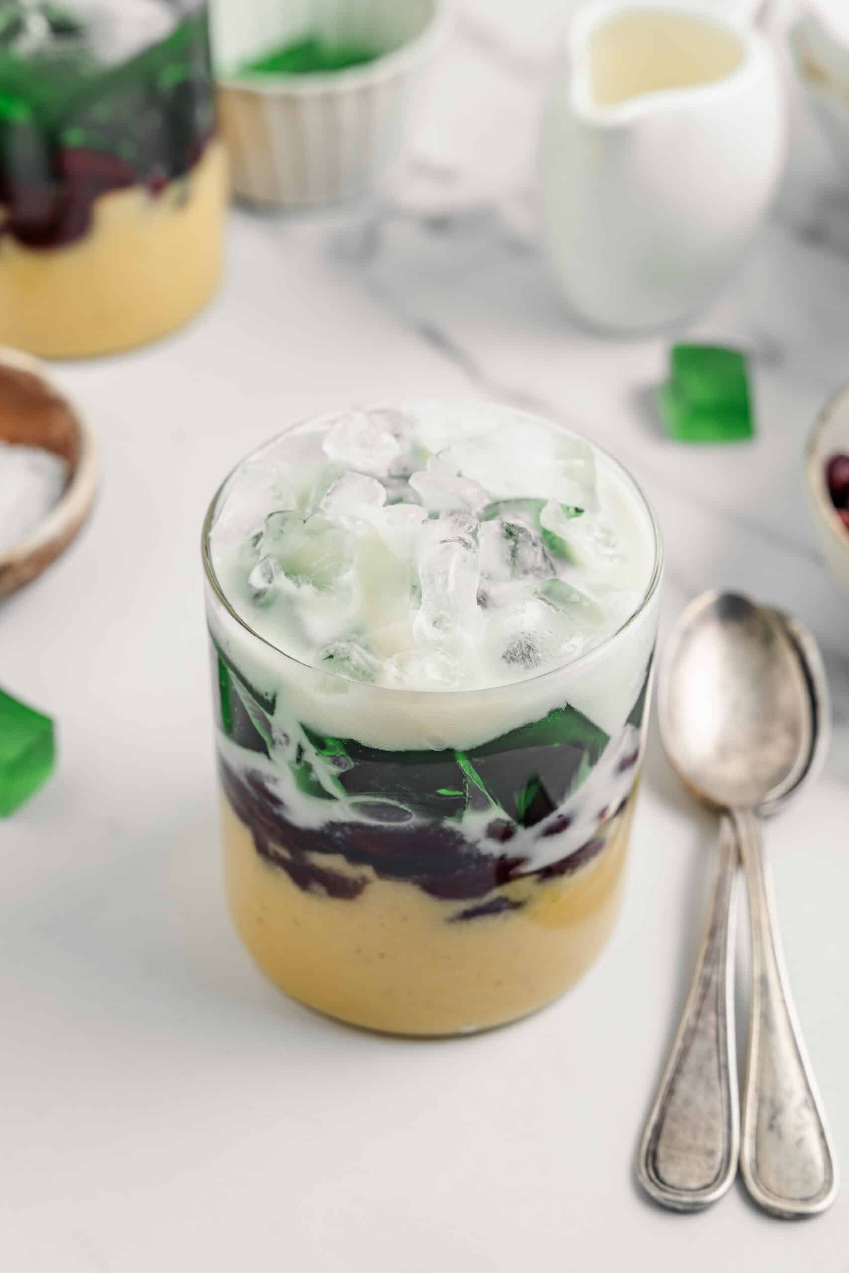 Che ba mau served in a clear glass with two spoons and bowls of toppings behind - Catherine Zhang Che ba mau served in a clear glass with two spoons and small bowls of toppings behind it
