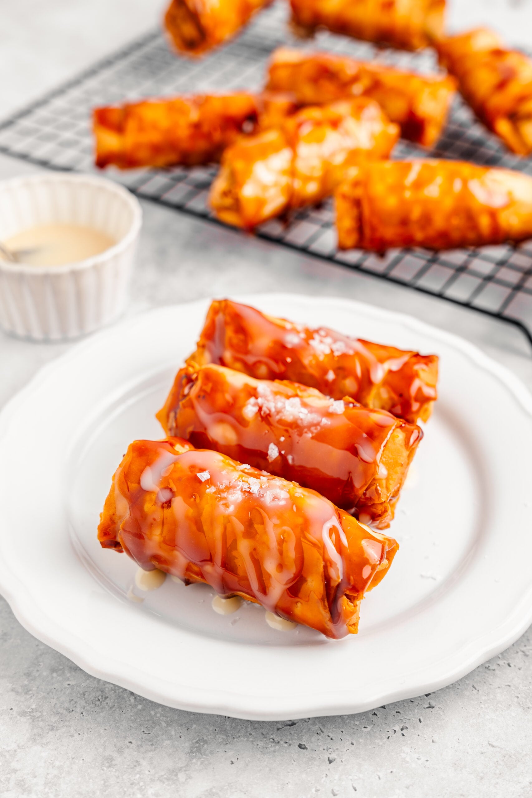 Three crispy turon Filipino banana lumpia on a white plate with condensed milk and flaky salt - Catherine Zhang Three crispy turon Filipino banana lumpia on a white plate with condensed milk and flaky salt