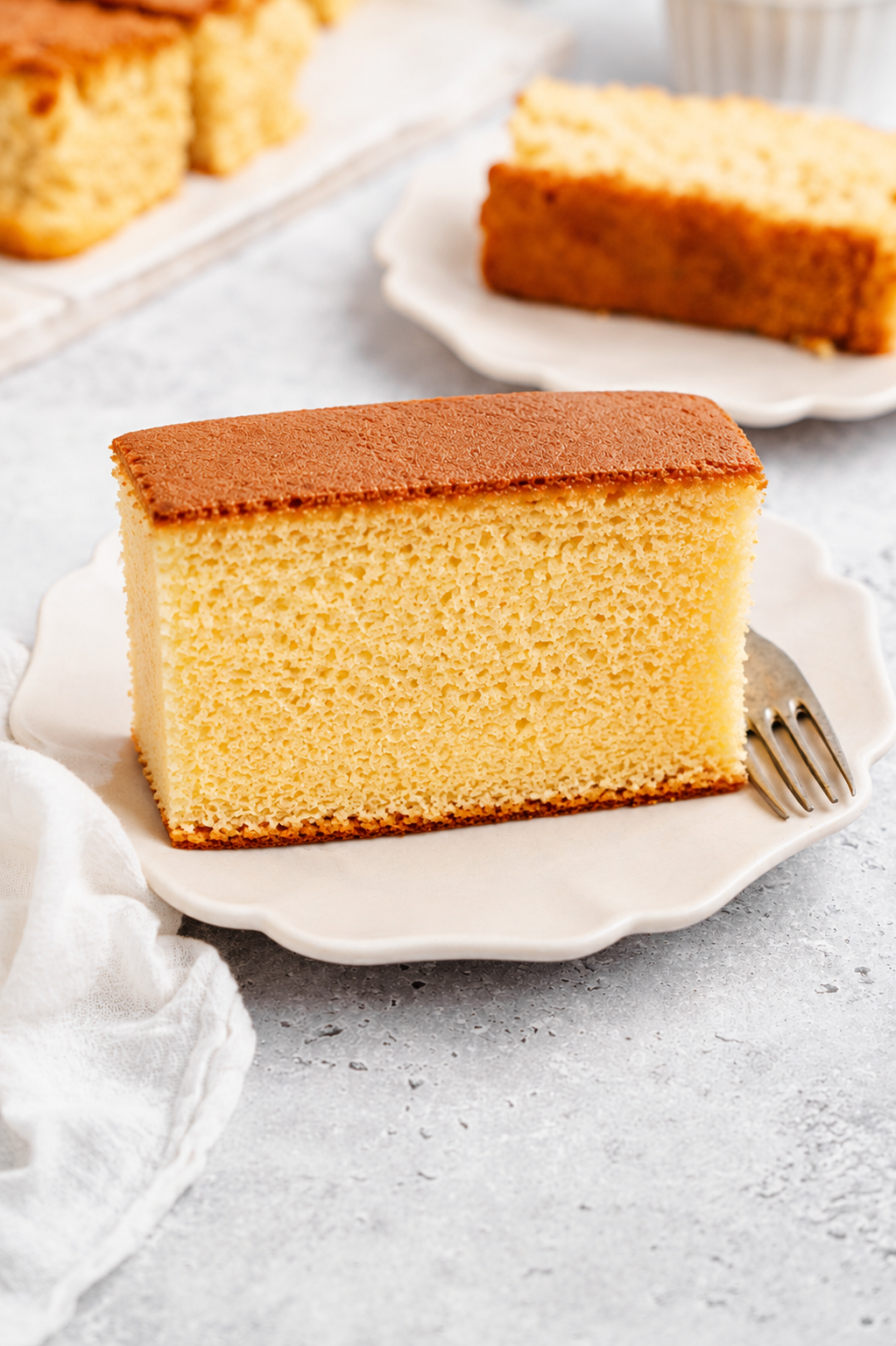 Thick slice of Japanese castella cake standing upright on a white plate with a dark honey crust on top - Catherine Zhang Thick slice of Japanese castella cake standing upright on a white plate with a dark honey crust on top