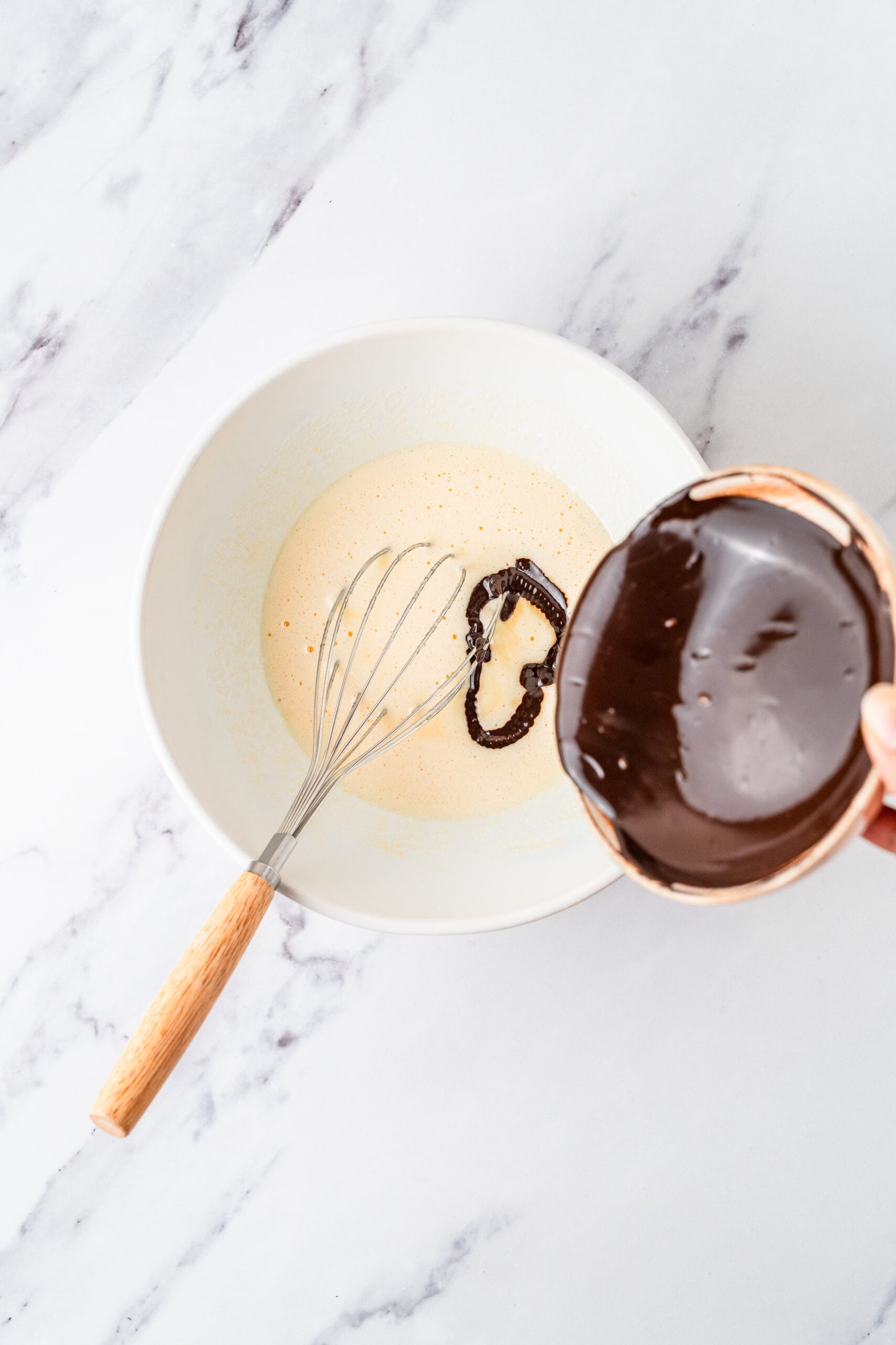 chocolate-crinkle-cookies-adding-melted-chocolate - Catherine Zhang Whisk in a bowl of batter on a marble countertop with a chocolate-dipped pastry being coated.
