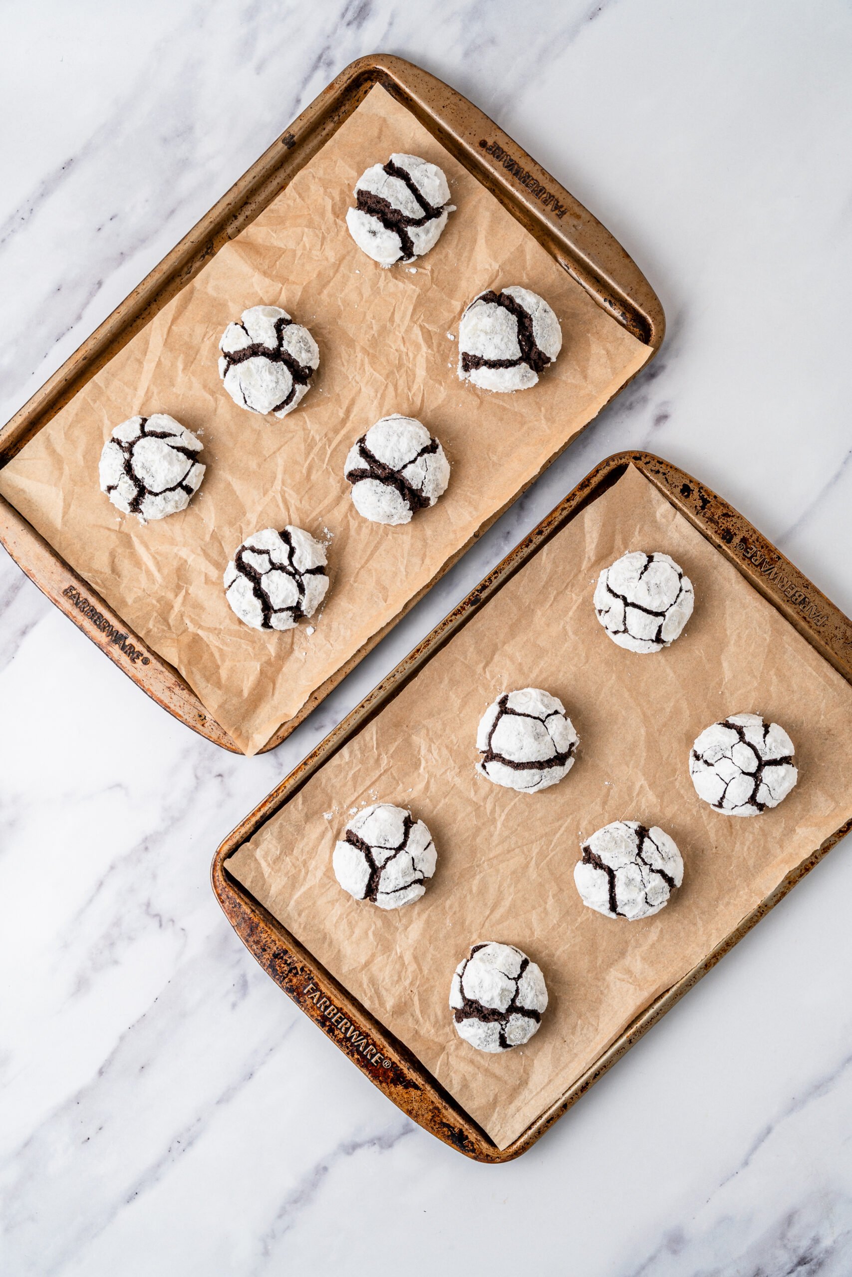 chocolate-crinkle-cookies-baked-crackle - Catherine Zhang Two baking sheets lined with parchment hold chocolate crinkle cookies dusted in powdered sugar on a marble surface