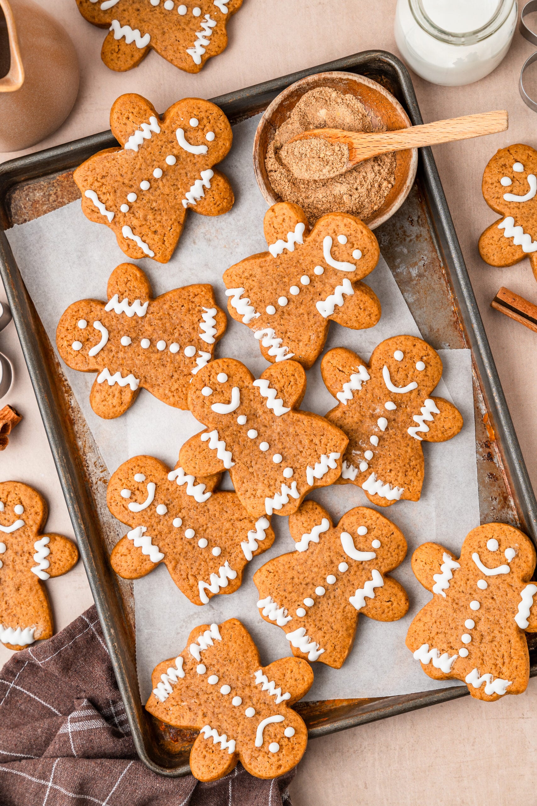 gingerbread-cookies-finished - Catherine Zhang Gingerbread men cookies with white icing on a parchment-lined baking sheet, with a wooden spoon and milk nearby.