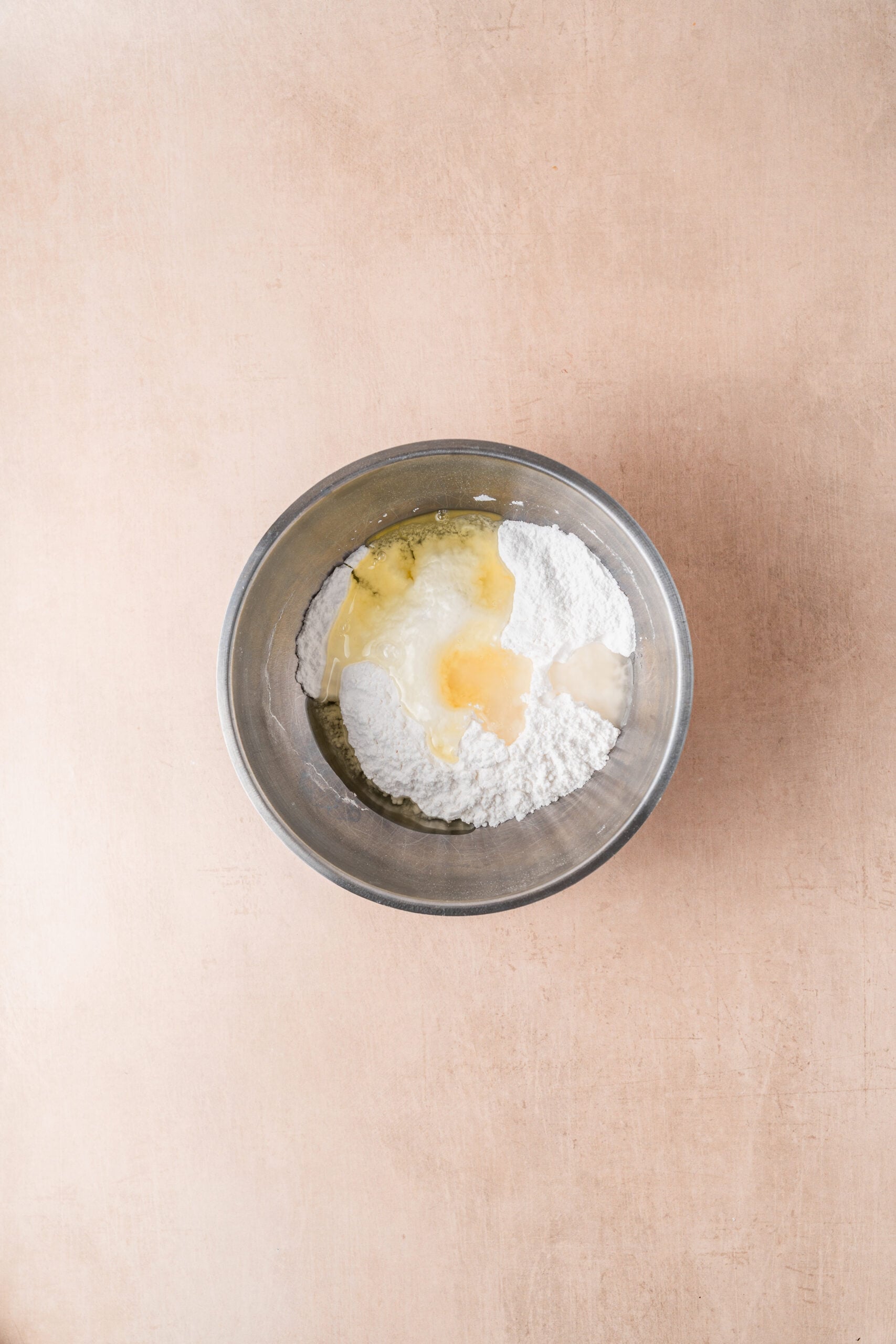 gingerbread-cookies-royal-icing-ingredients - Catherine Zhang Top-down view of a stainless steel mixing bowl containing flour and cracked eggs on a light brown surface