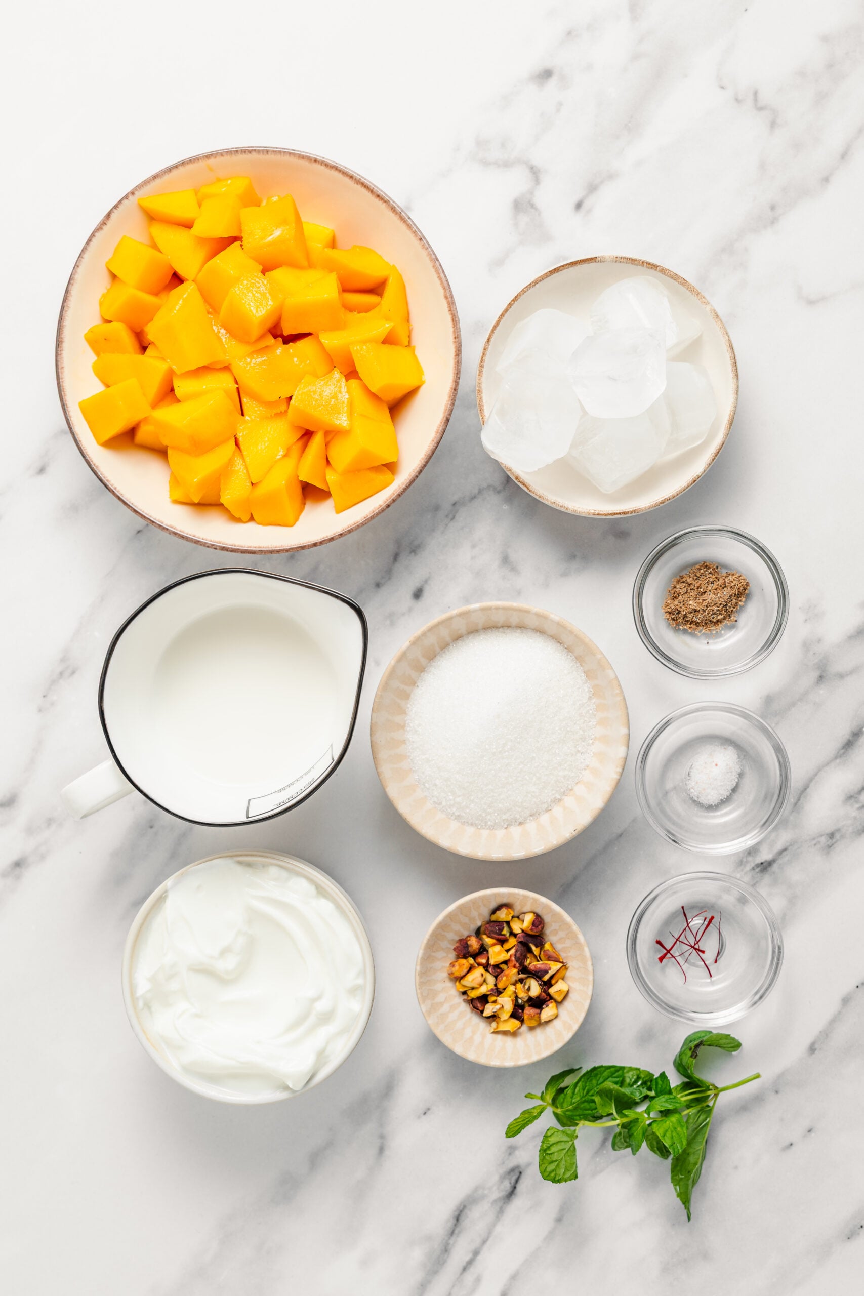 Mango lassi recipe ingredients - Catherine Zhang Ingredients for mango lassi on marble: diced mango, plain full fat yogurt, milk, sugar, ground cardamom, salt, pistachios and mint