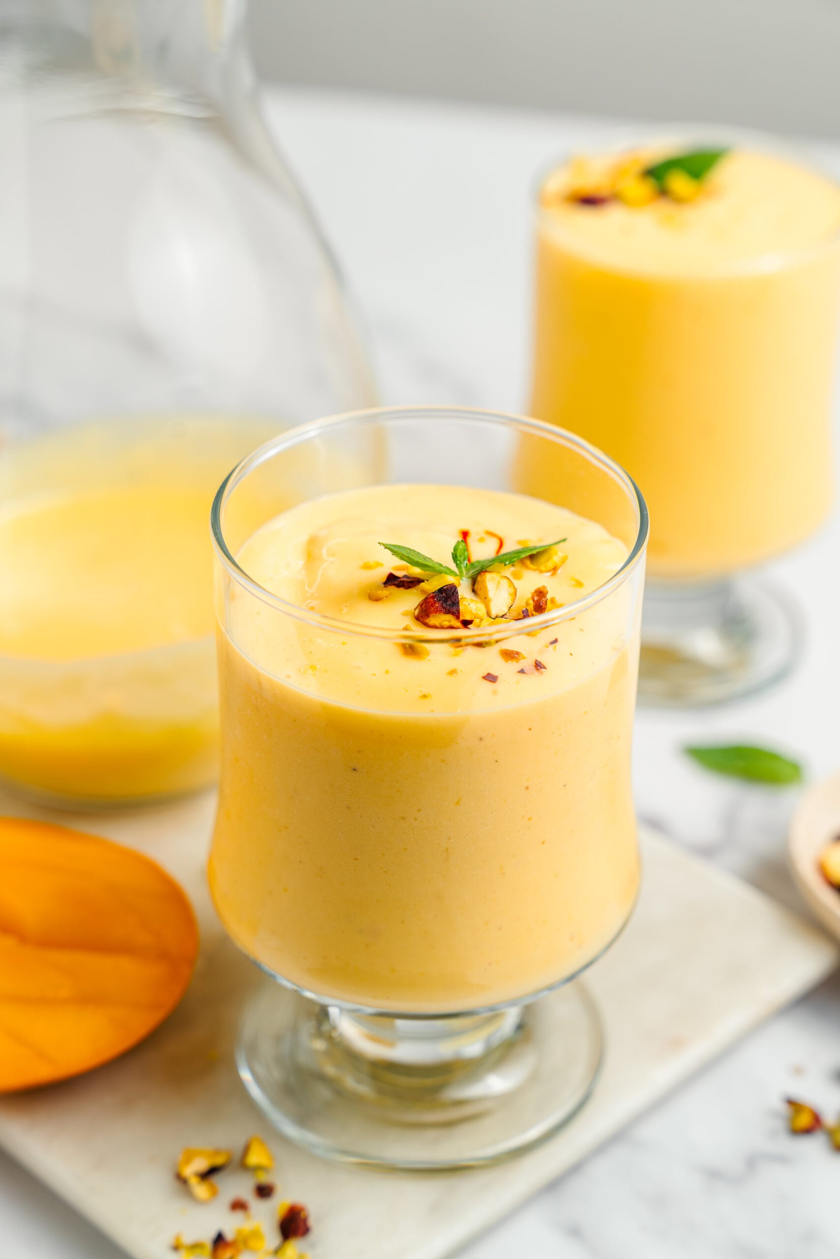 Mango lassi served in chilled glasses - Catherine Zhang Two glasses of thick mango lassi finished with chopped pistachios, saffron and mint