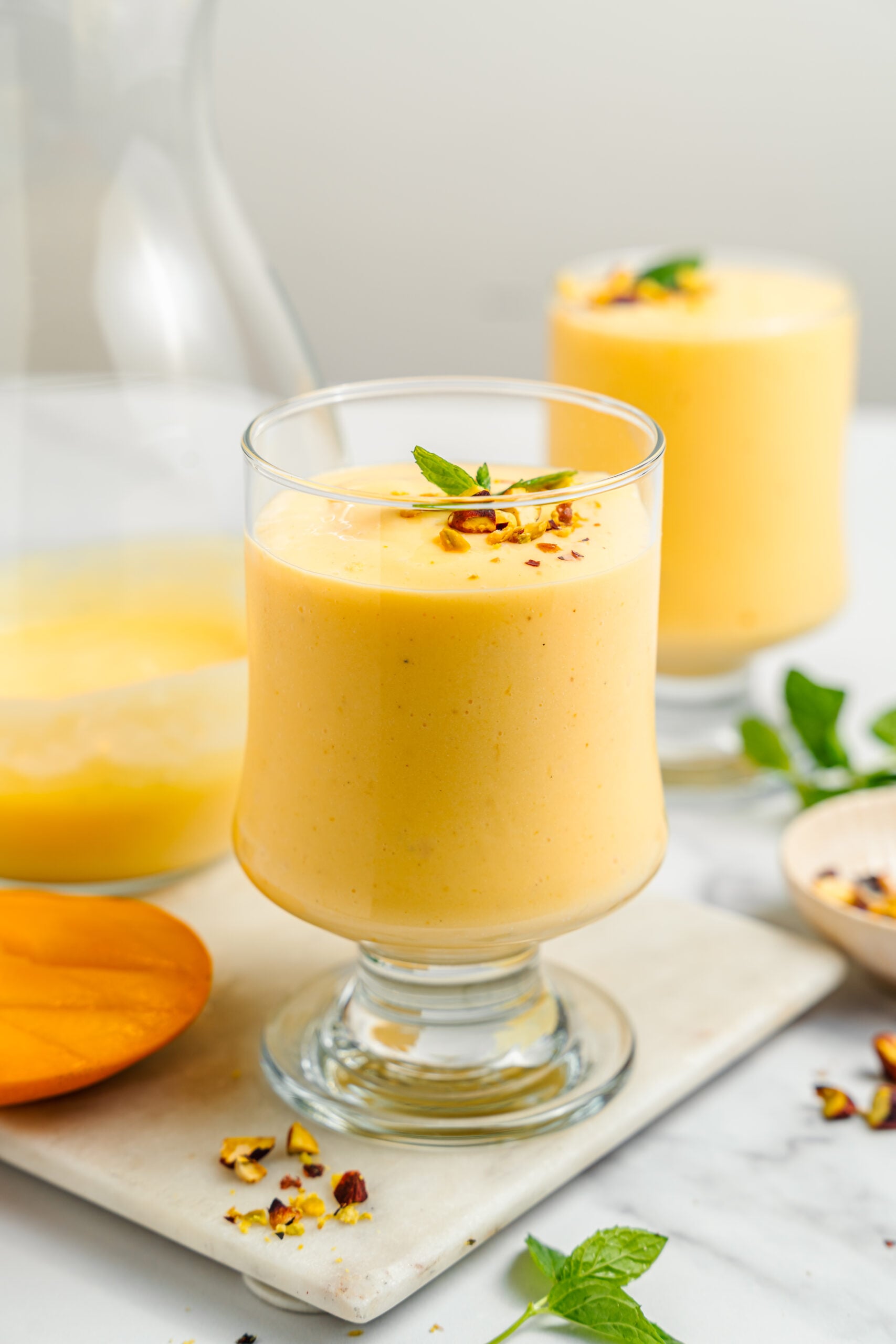 Two glasses of mango lassi - Catherine Zhang Two glasses of mango lassi on a marble board with sliced mango and scattered pistachios