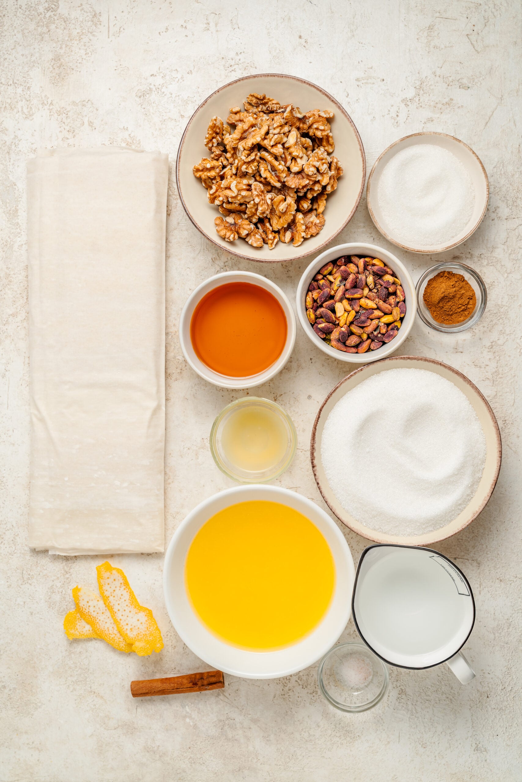 Pistachio baklava recipe ingredients - Catherine Zhang Ingredients for pistachio baklava laid out on a pale bench: phyllo pastry, walnuts, pistachios, honey, butter, sugar, cinnamon and lemon