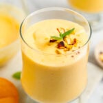 A tall glass of thick restaurant style mango lassi topped with chopped pistachios and mint