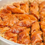 Close-up of golden baklava pieces in a baking dish, sprinkled with chopped nuts and sesame seeds, ready to serve